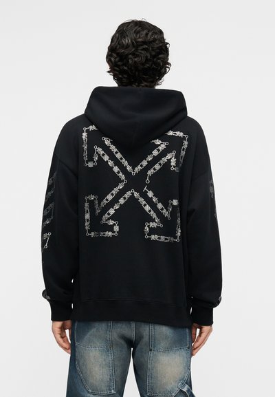 Persona ar cirtainiem tumšiem matiem valkā melnu kapuci ar lielu baltu bultiņas rakstu uz muguras un izbalējušas zilas džinsas, stāvot pret vienkāršu fonu.