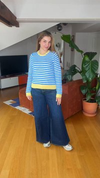 Jeune femme debout dans un salon, portant un pull rayé bleu et blanc avec des bords jaunes, un jean à jambes larges et des baskets argentées.