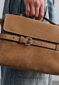 Mână care ține o geantă de umăr de la Moschino din suede bej, cu detalii de cataramă, purtată cu blugi din denim de un albastru deschis.