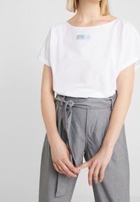 Vit bomull T-shirt med vid halsringning och korta ärmar, med ljusblå "DRYKORN"-text. Matchas med gråa tygbyxor med bälte.