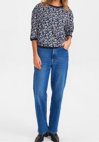 Frau in einer navyblauen, floralen Bluse und hochgeschnittenen blauen Jeans mit schwarzen Stiefeletten, die mit einer Hand in der Tasche steht.