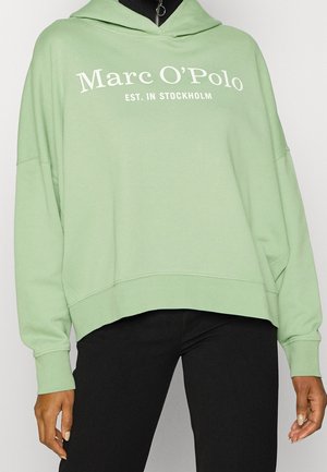 Person som bär en ljusgrön hoodie med texten "Marc O’Polo EST. I STOCKHOLM", kombinerad med svarta byxor, stående mot en neutral bakgrund.