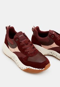 Baskets bordeaux en daim et mesh, avec une semelle intermédiaire blanche texturée parsemée de taches noires et un accent rose sur le côté.