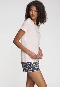 Lichtroze katoenen T-shirt met korte mouwen, voorzien van groene tekst, gecombineerd met zwarte shorts met bloemenprint met roze en witte bloemen.