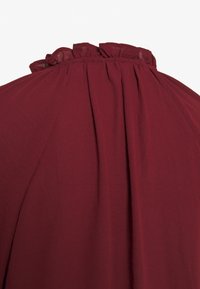 Abito bordeaux con colletto arricciato, texture morbida e tessuto arricciato all'altezza del collo, che crea una silhouette fluida.