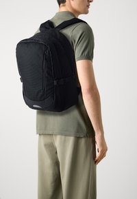 Mochila negra con un frente texturizado, correas ajustables y un diseño elegante. Llevada en la espalda de una persona con un atuendo casual.