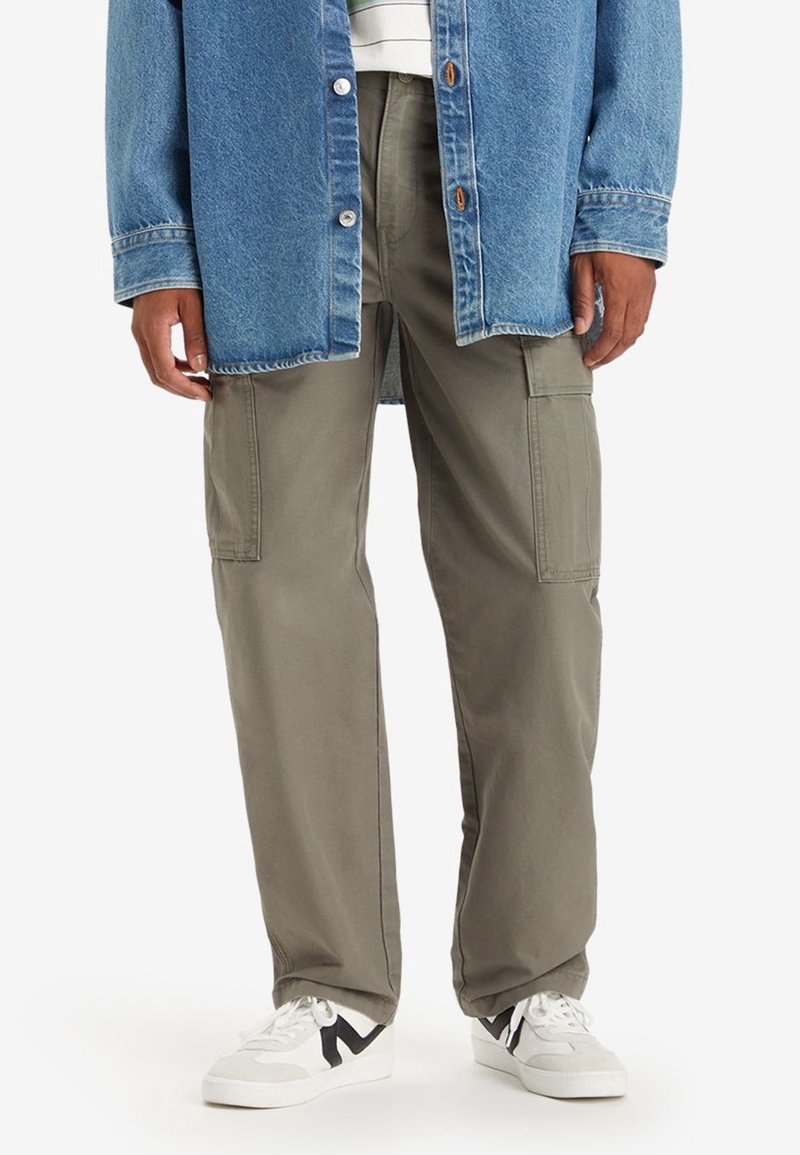 Gröna cargobyxor med sidofickor, matchat med en blå jeansjacka och vita sneakers med svarta detaljer. Avslappnad design.