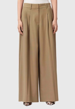 Kvinde iført beige bukser med høj talje og vide ben, kombineret med spidse dyreprintede sko, stående mod en grå baggrund.