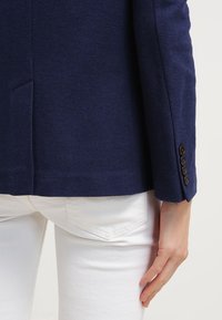 Blazer texturé bleu marine avec une coupe ajustée, fermeture à un bouton et trois boutons aux poignets, associé à un pantalon blanc.