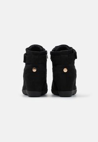 Ett par svarta mocka wedge sneakers sedd bakifrån, med vadderade kragar, ankelremmar med kardborre och små runda metallaccenter.