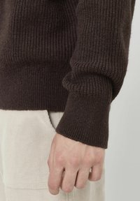 Pull en tricot marron avec texture côtelée, montrant le détail des poignets et un tissu texturé de couleur claire en dessous.