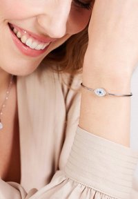Zilveren armband met een ovale hanger, afgebeeld met een ontwerp van een blauwe oog en meerdere kleine heldere edelstenen. De pols is gedeeltelijk zichtbaar en draagt een lichtgekleurde blouse met gestructureerde mouwen.