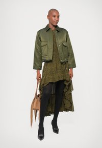 Oliivroheline lühike bomber-jakk kihilise tumerohelise mustriga kleidi peal, milles on mustad sukkpüksid ja terava ninaga kontsad, hoides käes frändide jaotusega kotti.