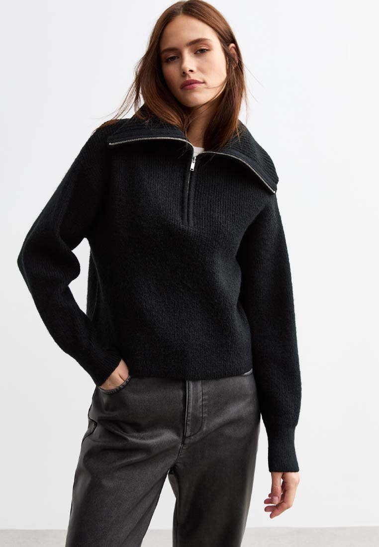 Svart stickad tröja med hög krage och halvdragkedja; har en strukturerad tyg och en avslappnad passform, kombinerad med mörka läderbyxor.