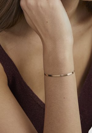 Frau trägt ein dünnes goldenes Armband am Handgelenk, die Hand ruht nahe am Gesicht, gekleidet in einem dunkelvioletten ärmellosen Oberteil.