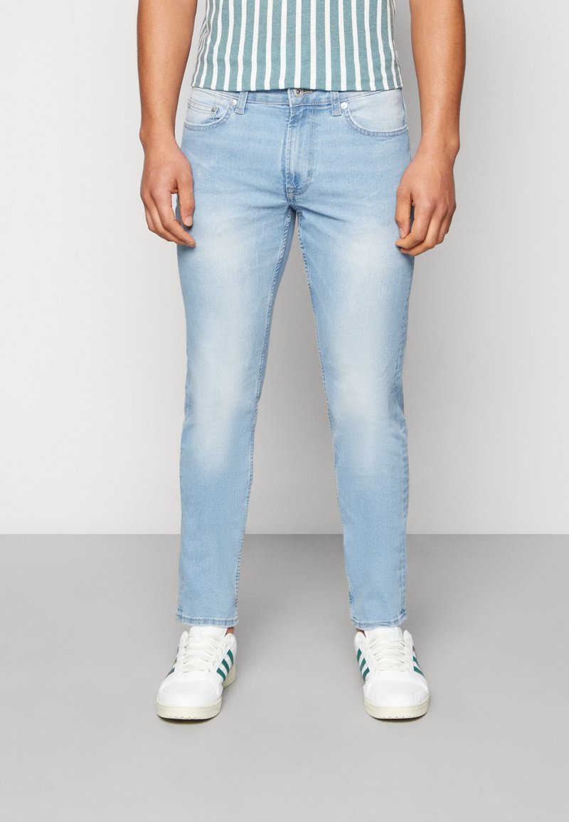 Homme portant un jean slim bleu clair et des baskets blanches avec des rayures vertes, se tenant devant un fond clair uni.