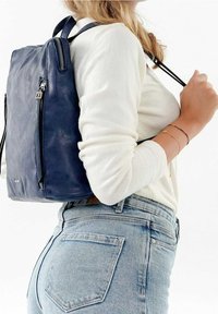 Námořnický kožený batoh s texturovaným povrchem, s přední kapsou na zip a dvojitými ramenními popruhy, nošený na rameni osoby.