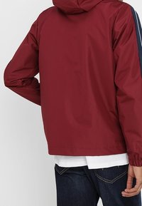 Chaqueta con capucha en color burdeos, de textura suave y brillante; presenta franjas laterales en color azul marino. Diseño simple, de longitud corta, con puños y dobladillo elásticos.