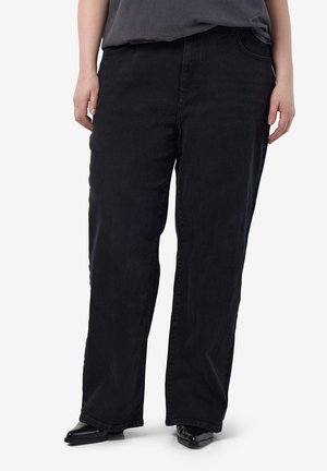 Pessoa a usar calças pretas largas com bolsos frontais, combinadas com sapatos pretos brilhantes de bico fino e uma parte de cima escura, de pé contra um fundo claro.