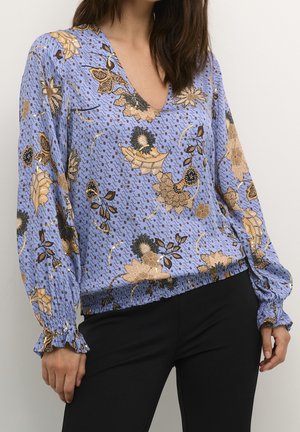 Kobieta ubrana w niebieską, kwiecistą bluzkę z długim rękawem z falbankami na mankietach oraz czarne spodnie, stojąca na jednolitym tle.