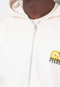 Primo piano di una persona che indossa una felpa bianca con zip e una grafica di limone con il testo "PEGAD" sul petto.