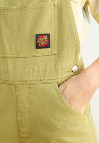 La salopette in cotone verde oliva presenta una tasca frontale, una patch con logo e bottoni a pressione metallici. Il tessuto mostra una finitura strutturata.