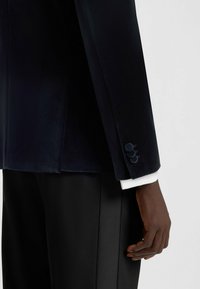 Blazer in velluto blu navy con tre bottoni decorativi sui polsini, abbinato a pantaloni neri su misura, caratterizzati da una texture liscia e morbida.