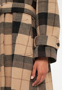 Manteau en laine à carreaux beige et noir avec une taille ceinturée, présentant un poignet replié et une poche latérale. La texture est douce et à motifs.