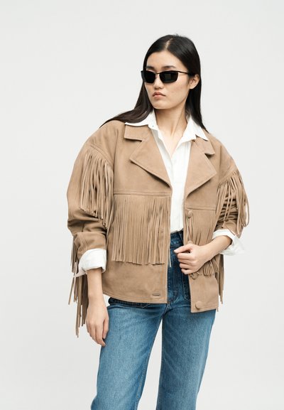 Vrouw met zwarte zonnebril, beige suède jas met franjes, wit shirt en blauwe spijkerbroek, staand tegen een effen lichte achtergrond.