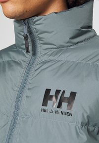 Szara pikowana kurtka z wysokim kołnierzem, z czarnym logo "HH" i pełnym zamkiem błyskawicznym. Gładkie wykończenie z poziomymi przeszyciami.