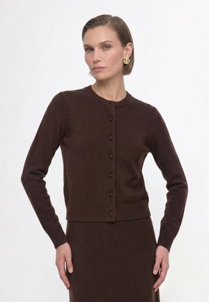 Woman with short brown hair wearing gold earrings and a dark brown buttoned cardigan over a matching skirt against a plain white background.