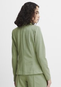 Green fitted blazer with textured fabric, stand collar, and tailored seams. Features a simple design and matching green outfit below.