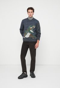 Pull en tricot avec un motif de dinosaure en vert, marron et blanc sur fond bleu foncé, associé à un pantalon en velours côtelé noir et des baskets noires.