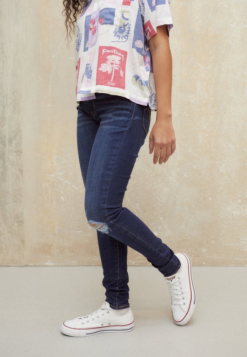 Personne portant un jean skinny déchiré bleu foncé, des baskets blanches avec des rayures rouges et bleues, et une chemise à motifs colorés, debout sur un sol clair.