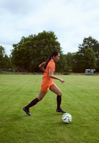 Oranžový futbalový dres s čiernymi detailmi, čierne ponožky po kolená a čierne kopačky. Hráč driblovanie s viacfarebnou futbalovou loptou na trávnatom ihrisku.