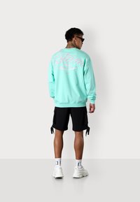 Mint green sweatshirt with a large pink graphic on the back, paired with black cargo shorts, and white sneakers with black accents.