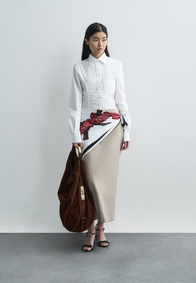 Γυναίκα με λευκό ριχτό πουκάμισο, μπεζ φούστα με λεπτομέρεια από φουλάρι, μαύρες σαγιονάρες Moschino και μεγάλη καφέ τσάντα Moschino, που στέκεται μπροστά σε έναν απλό τοίχο.