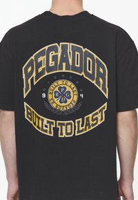 Černé bavlněné tričko se velkým tištěným textem v žluté a bílé barvě: "PEGADOR" a "BUILT TO LAST" s emblémem a dekorativními akcenty.