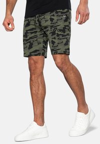 Pantaloni corti mimetici in verde scuro e nero, realizzati in tessuto morbido. Presentano una vita con laccetto e tasche laterali, abbinati a sneaker bianche.