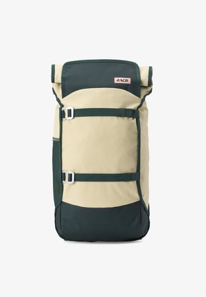 Sac à dos beige et vert foncé à rabat enroulable avec deux bretelles réglables à l'avant et une petite étiquette rose près du centre supérieur.