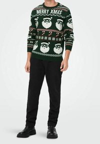 Grüner Weihnachtspullover mit weißen Santa-Gesichtern, Zuckerstangen und dem Text "MERRY XMAS". Gerippter Ausschnitt und Bündchen, kombiniert mit schwarzen Hosen und Schuhen.