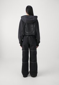 Vrouw met lang zwart haar, gekleed in een zwarte cropped hoodie met een smiley-gezicht en zwarte wijde broek met witte stiksels, staande tegen een neutrale achtergrond.