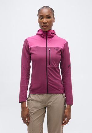 Vrouw draagt een aangesloten tweekleurig magentakleurig vest met rits en capuchon en beige broek, staand tegen een effen lichte achtergrond.