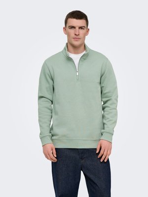Homme portant un sweat à col zippé vert clair sur un t-shirt blanc et un jean bleu foncé, face à l'avant avec une expression neutre.