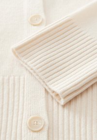 Nahaufnahme eines cremefarbigen, gerippten Strickcardigans mit einer Ärmel- und geknöpften Vorderseite sowie runden, cremefarbenen Knöpfen.