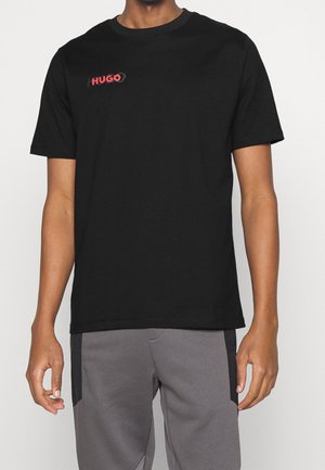 Homme portant un t-shirt noir à manches courtes avec le logo rouge "HUGO" sur la poitrine gauche et un pantalon gris avec des panneaux latéraux noirs.