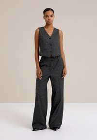 Una modelo se presenta con confianza en un chaleco gris a medida y pantalones de pierna ancha, con un fondo neutral.