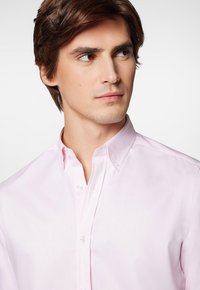 Jasnor�óżowa koszula zapinana na guziki z klasycznym kołnierzykiem i perłowymi guzikami. Gładka tekstura materiału, dopasowany krój i brak widocznych wzorów.