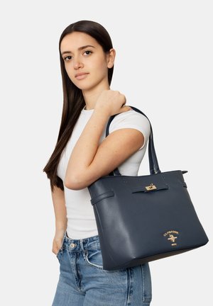 Junge Frau mit langen dunklen Haaren trägt ein weißes T-Shirt und Jeans und trägt eine dunkelblaue Lederhandtasche über der Schulter.