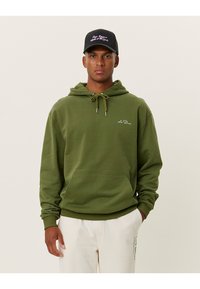 Felpa verde oliva in morbido tessuto con tasca a marsupio frontale e cappuccio regolabile con cordini. Abbinata a un cappellino nero con ricami rosa.